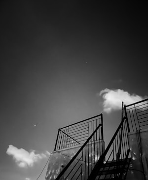 A black and white image showcasing a metal staircase with railings leading upward, set against a backdrop of a slightly cloudy sky. The perspective is angled from below the staircase, giving a sense of ascent and space.