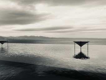 A serene, monochrome landscape featuring two symmetrical triangular structures rising from a still body of water. The scene is set against a backdrop of distant mountains under a cloudy sky, creating a calm and minimalist atmosphere.