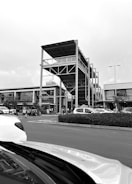 A busy parking lot in front of a modern shopping complex featuring a uniquely designed multi-level structure with open sides and a roof. Cars are parked while several people are walking around the area. The sky is overcast, adding a monochromatic tone to the scene.