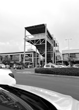 A busy parking lot in front of a modern shopping complex featuring a uniquely designed multi-level structure with open sides and a roof. Cars are parked while several people are walking around the area. The sky is overcast, adding a monochromatic tone to the scene.