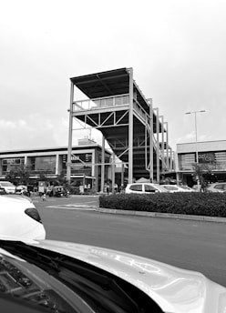 A busy parking lot in front of a modern shopping complex featuring a uniquely designed multi-level structure with open sides and a roof. Cars are parked while several people are walking around the area. The sky is overcast, adding a monochromatic tone to the scene.