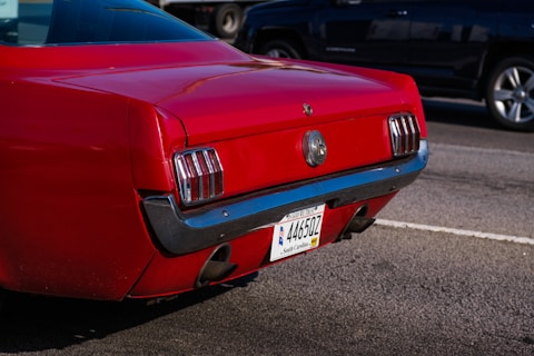A close-up view of the rear section of a red classic car, featuring its distinct tail lights and chrome bumper. The license plate identifies the car as registered in South Carolina. The shiny red paint contrasts with the asphalt road surface and other vehicles in the background.