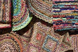 An array of colorful, woven, braided rugs featuring intricate patterns and vibrant textiles. The rugs display a variety of geometric designs, including circular and rectangular shapes, with a mix of bright colors like red, blue, green, yellow, and pink. The weaving technique is evident, showcasing diverse textures and craftsmanship.