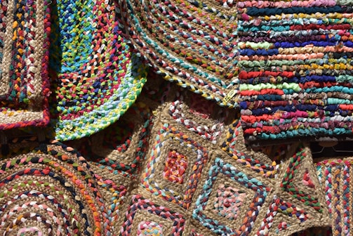 An array of colorful, woven, braided rugs featuring intricate patterns and vibrant textiles. The rugs display a variety of geometric designs, including circular and rectangular shapes, with a mix of bright colors like red, blue, green, yellow, and pink. The weaving technique is evident, showcasing diverse textures and craftsmanship.