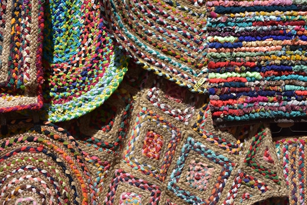 An array of colorful, woven, braided rugs featuring intricate patterns and vibrant textiles. The rugs display a variety of geometric designs, including circular and rectangular shapes, with a mix of bright colors like red, blue, green, yellow, and pink. The weaving technique is evident, showcasing diverse textures and craftsmanship.