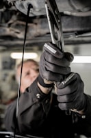 A close-up view of a person using pliers to work on machinery, with oil visibly draining from the machinery above. The individual is wearing protective gloves and a uniform. The setting appears to be an industrial environment, likely a workshop or garage.