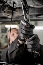 A close-up view of a person using pliers to work on machinery, with oil visibly draining from the machinery above. The individual is wearing protective gloves and a uniform. The setting appears to be an industrial environment, likely a workshop or garage.