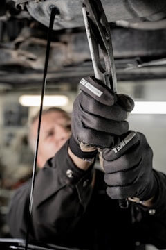A close-up view of a person using pliers to work on machinery, with oil visibly draining from the machinery above. The individual is wearing protective gloves and a uniform. The setting appears to be an industrial environment, likely a workshop or garage.