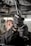 A close-up view of a person using pliers to work on machinery, with oil visibly draining from the machinery above. The individual is wearing protective gloves and a uniform. The setting appears to be an industrial environment, likely a workshop or garage.