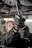 A close-up view of a person using pliers to work on machinery, with oil visibly draining from the machinery above. The individual is wearing protective gloves and a uniform. The setting appears to be an industrial environment, likely a workshop or garage.