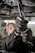 A close-up view of a person using pliers to work on machinery, with oil visibly draining from the machinery above. The individual is wearing protective gloves and a uniform. The setting appears to be an industrial environment, likely a workshop or garage.