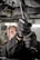 A close-up view of a person using pliers to work on machinery, with oil visibly draining from the machinery above. The individual is wearing protective gloves and a uniform. The setting appears to be an industrial environment, likely a workshop or garage.