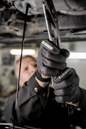 A close-up view of a person using pliers to work on machinery, with oil visibly draining from the machinery above. The individual is wearing protective gloves and a uniform. The setting appears to be an industrial environment, likely a workshop or garage.