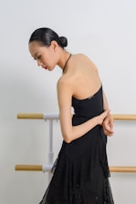 A woman with a sleek hairstyle is elegantly posed in a black dress, standing beside wooden ballet barres against a plain backdrop.