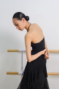 A woman with a sleek hairstyle is elegantly posed in a black dress, standing beside wooden ballet barres against a plain backdrop.
