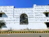 A white building facade with an archway in the center. The wall is adorned with text in bold letters. There are shadows cast by sunlight, and the sky is clear and blue. A small tree is visible on the left, near a sidewalk and a road with a yellow and black painted curb.