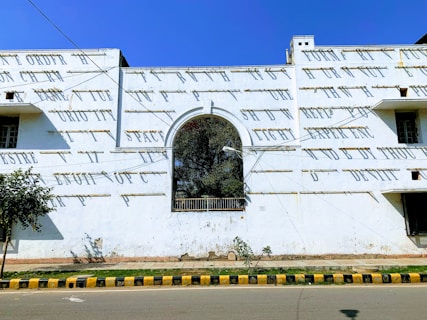 A white building facade with an archway in the center. The wall is adorned with text in bold letters. There are shadows cast by sunlight, and the sky is clear and blue. A small tree is visible on the left, near a sidewalk and a road with a yellow and black painted curb.