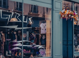 A storefront with large glass windows displays various clothing items, including mannequins wearing colorful outfits and accessories. The reflection of nearby buildings and a parked car is visible in the glass. Above the entrance, a vibrant arrangement of flowers adds a touch of color to the exterior.