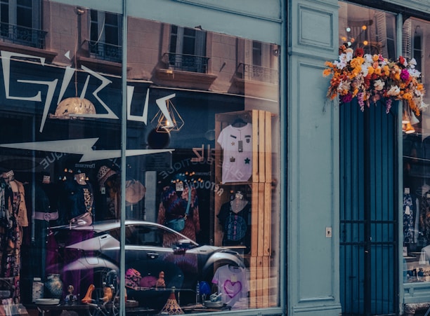 A storefront with large glass windows displays various clothing items, including mannequins wearing colorful outfits and accessories. The reflection of nearby buildings and a parked car is visible in the glass. Above the entrance, a vibrant arrangement of flowers adds a touch of color to the exterior.