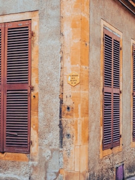 A corner of an old building with textured walls displaying a worn appearance. Three wooden shutters painted in a reddish-brown color cover the windows. A small sign with French text is affixed to the stone wall.