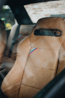 A close-up view of a tan leather car seat with a racing stripe detail, seen through a car window reflecting some outdoor scenery. The setting appears to be inside a parked car, with a blurred background visible through the window.