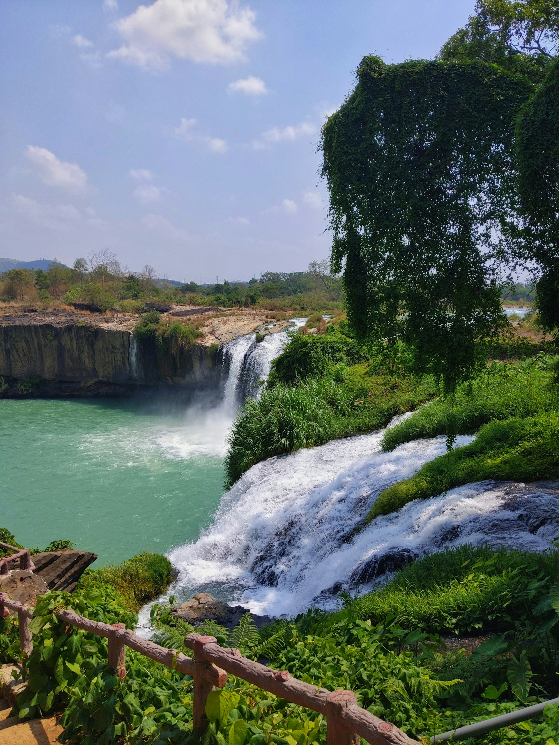 A stunning waterfall cascading into a crystal-clear pool, surrounded by vibrant tropical foliage under a bright sky.