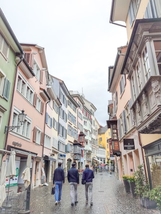 A narrow cobblestone street lined with colorful, historical buildings featuring decorative facades and shuttered windows. Some people are walking along the street, and there are small outdoor seating areas with umbrellas. The scene suggests a quaint, possibly European town.
