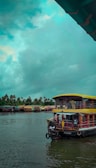 A traditional houseboat is floating on a serene body of water under a cloudy sky. The boat has a unique design with a thatched roof and is adorned with vibrant colors. In the background, more houseboats line the water's edge, surrounded by lush palm trees.