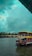 A traditional houseboat is floating on a serene body of water under a cloudy sky. The boat has a unique design with a thatched roof and is adorned with vibrant colors. In the background, more houseboats line the water's edge, surrounded by lush palm trees.