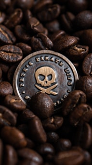 A metal coin with a skull and crossbones design is surrounded by roasted coffee beans. The coin features an engraved text and a detailed pirate emblem, while the coffee beans display a rich texture and a deep brown color.