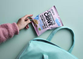 A hand with green-painted nails holds a bag of organic protein chips labeled 'The Organic Crave' against a light teal background. The person is wearing a pink fuzzy sweater. A teal mesh bag is also visible in the image.