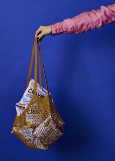 A hand wearing a pink sleeve holds a net-style grocery bag filled with assorted packaged food products against a solid blue background.