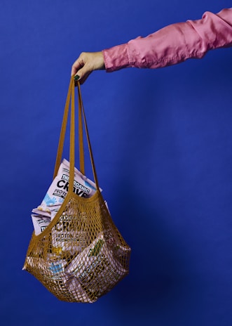 A hand wearing a pink sleeve holds a net-style grocery bag filled with assorted packaged food products against a solid blue background.