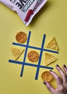 A tic-tac-toe game is set up with tortilla chips acting as the game pieces on a yellow background. Circular chips and triangular chips are in play on a grid made from blue tape. A hand with painted nails is about to place a circular chip on the grid. A bag of chips is partially visible at the top.