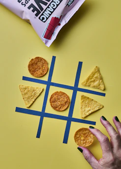 A tic-tac-toe game is set up with tortilla chips acting as the game pieces on a yellow background. Circular chips and triangular chips are in play on a grid made from blue tape. A hand with painted nails is about to place a circular chip on the grid. A bag of chips is partially visible at the top.