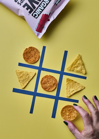 A tic-tac-toe game is set up with tortilla chips acting as the game pieces on a yellow background. Circular chips and triangular chips are in play on a grid made from blue tape. A hand with painted nails is about to place a circular chip on the grid. A bag of chips is partially visible at the top.