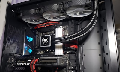 A high-performance gaming computer setup showing the interior components, including a GeForce RTX graphics card, cooling system with three fans, a motherboard, and visible wiring with red and black cables.