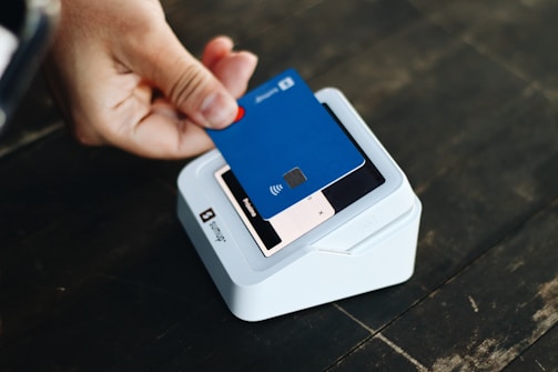 A hand is holding a blue credit card above a white card reader placed on a dark surface. The card features contactless payment symbols, and the reader is positioned on a wooden table.