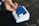A hand is holding a blue credit card above a white card reader placed on a dark surface. The card features contactless payment symbols, and the reader is positioned on a wooden table.