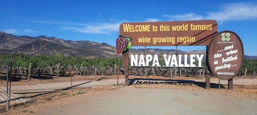A large wooden sign welcomes visitors to Napa Valley, known for its world-famous wine-growing region. The sign includes illustrations of grapes and vineyards, along with a quote about wine. Behind the sign, rows of grapevines stretch towards rolling hills under a clear blue sky.