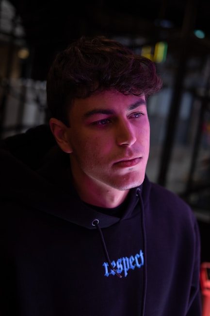 A young person with brown hair is wearing a black hoodie with the word 'respect' in blue lettering. The face is illuminated with pink and purple lighting, giving a neon effect. The background is blurred, with dark and muted colors.