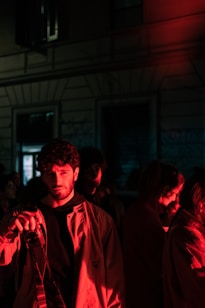 A group of people stand in dim lighting with a strong red hue casting over them. One individual in the foreground is holding a camera and looking ahead, partially illuminated by the red light. The background is dark and indistinct, suggesting a nighttime setting or an indoor environment with minimal lighting.