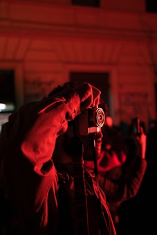 A person is holding a camera up to their eye, partially illuminated by red ambient light. The background is blurred, suggesting an urban environment with graffiti on walls. The scene captures a moment during night time, characterized by the dominant presence of red and dark shadows.