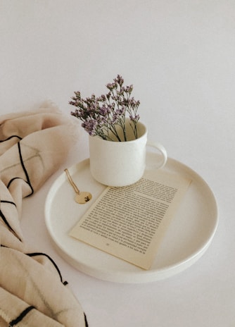 A soft monochrome mug placed on a linen cloth next to an open book and a small vase of dried flowers.