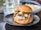 A gourmet burger sits on a black plate, featuring a toasted bun topped with poppy seeds, melted cheese, pickles, and caramelized onions. The burger appears juicy with cheese oozing down the sides. In the background, there's a glass and a blurred bowl of fries.