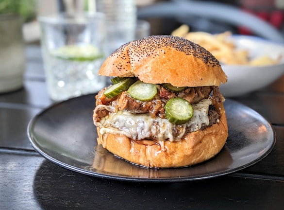 A gourmet burger sits on a black plate, featuring a toasted bun topped with poppy seeds, melted cheese, pickles, and caramelized onions. The burger appears juicy with cheese oozing down the sides. In the background, there's a glass and a blurred bowl of fries.