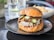 A gourmet burger sits on a black plate, featuring a toasted bun topped with poppy seeds, melted cheese, pickles, and caramelized onions. The burger appears juicy with cheese oozing down the sides. In the background, there's a glass and a blurred bowl of fries.