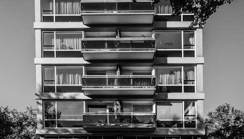 A black and white photograph of a modern multi-story apartment building with large glass windows and balconies. Each balcony has a railing, and some windows have curtains or blinds. The shadows of trees are visible, and there is a tree branch partially visible in the upper right corner.
