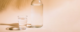 A clear glass and a transparent bottle, both filled with water, are placed on a light beige background. The lighting creates subtle shadows, highlighting the simplicity and clarity of the objects.