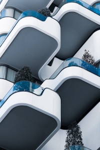 A series of modern, curved balconies with blue-tinted glass railings are stacked vertically. The architectural design is sleek, with white and gray tones dominating the structure. Each balcony has a decorative plant, adding a touch of greenery.